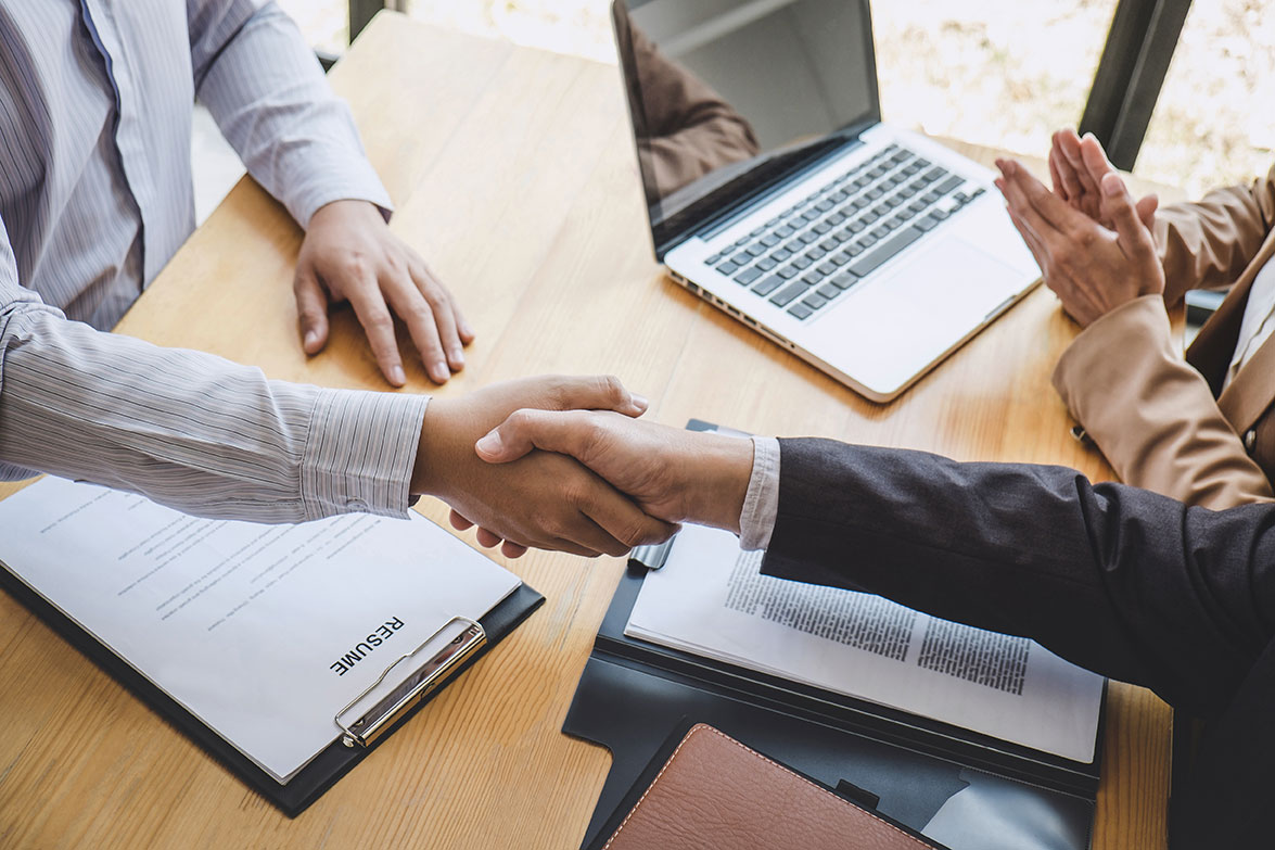 Two people shake hands across a table while a third person claps.